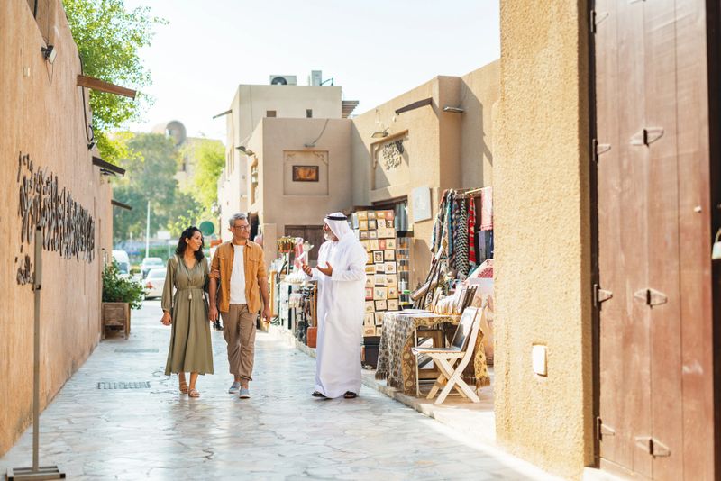 A senior Arabic tour guide leads a mid-adult couple through a sunlit historic Old Dubai alley featuring traditional architecture, shops, and vibrant cultural details. The warm, golden lighting enhances the charm of the bustling outdoor setting.