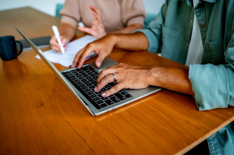 Two colleagues working together at a wooden desk, focusing on a laptop. Shows collaboration and productivity in a professional setting. Ideal for business and office themes highlighting teamwork and technology.