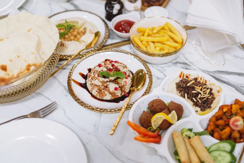A vibrant spread of Middle Eastern dishes featuring hummus, falafel, pita bread, fries, and garnished plates served on elegant golden tableware over a marble surface.
