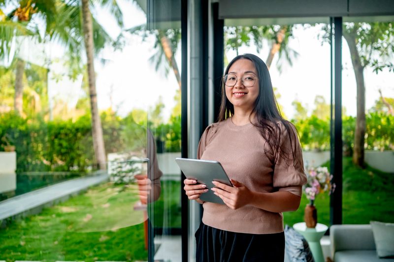 A young woman stands smiling, holding a tablet in a modern setting with lush greenery outside. The natural light filters through, creating a bright and inviting atmosphere.