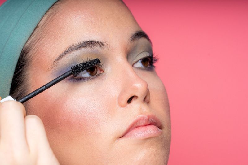 Close-up of a young woman applying mascara, enhancing her eyelashes with precision against a vibrant pink backdrop
