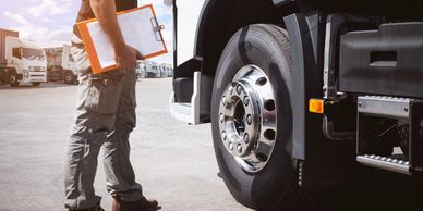 Worker inspecting a truck tire with a clipboard in hand at a truck yard.