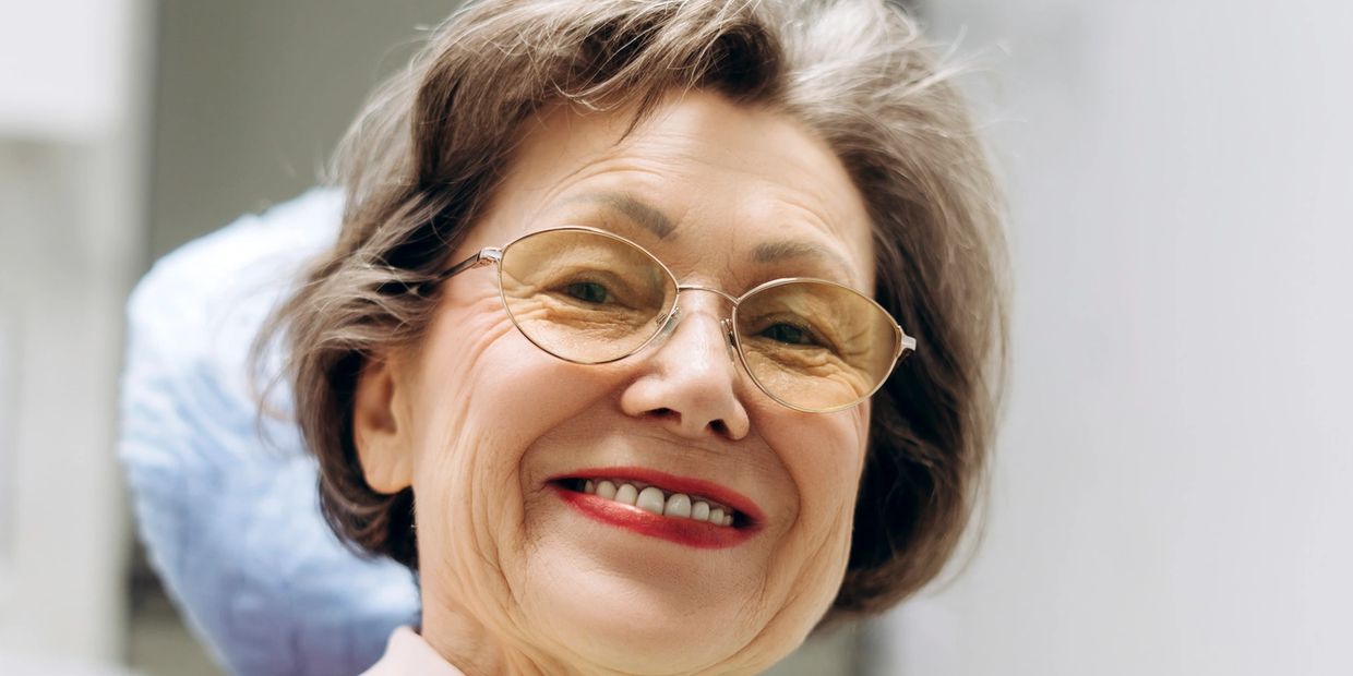 Smiling elderly woman wearing glasses with light brown hair and red lipstick.