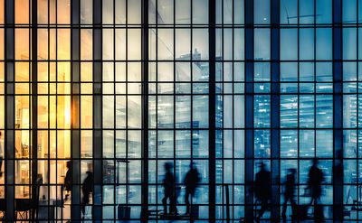 Silhouettes of people walking behind a large glass window with warm and cool lighting.
