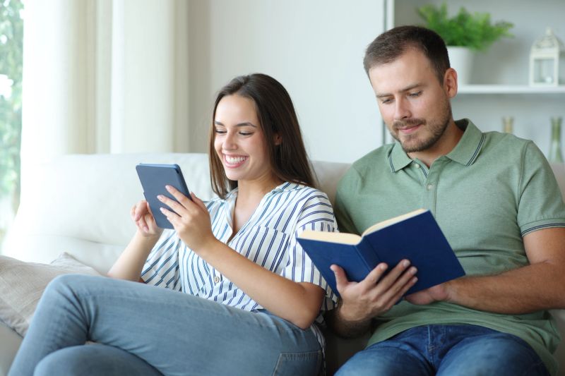 Couple reading a paper book and ebook sitting on a couch at home.