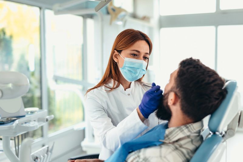 Female dentist, wearing a mask and gloves, examining male patient's teeth in a dental office, delivering expert oral care and promoting healthy dental hygiene practices