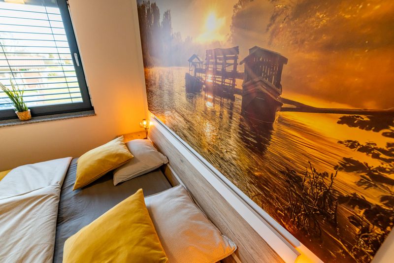 Close-Up of a Cozy Bedroom Interior Featuring a Warmly Illuminated Bedside Lamp,Golden Pillows,and a Striking Large-Format Wall Art of a Wooden Pier on a Sunlit River. Natural Light From a Nearby Window Enhances the Tranquil Atmosphere,with a Potted Plant on the Windowsill Adding a Touch of Greenery