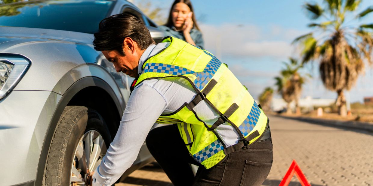 Man changing a car tire while woman talks on phone.