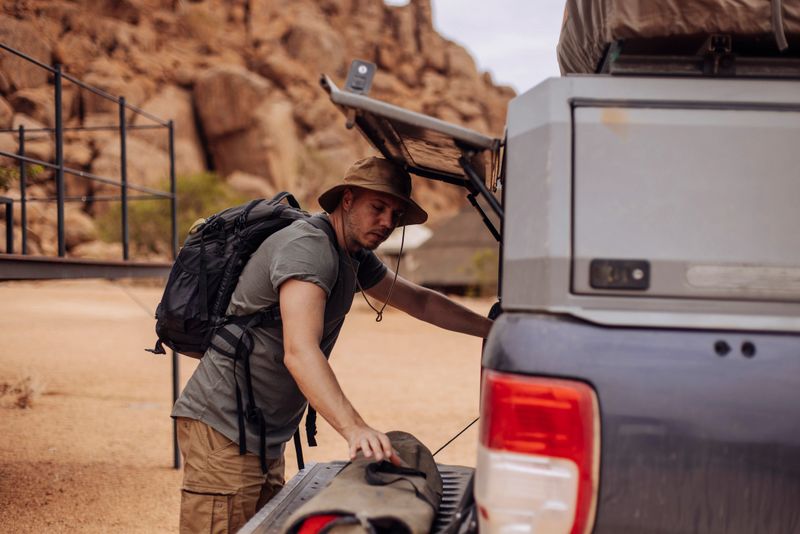 A man steps out of a safari lodge and places his black suitcase into a safari jeep, ready to continue his adventurous journey into the wild.