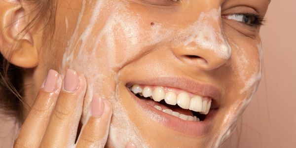 Smiling woman gently applying foamy facial cleanser to her cheek.
