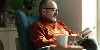 Man in orange sweater reading a book by the window.