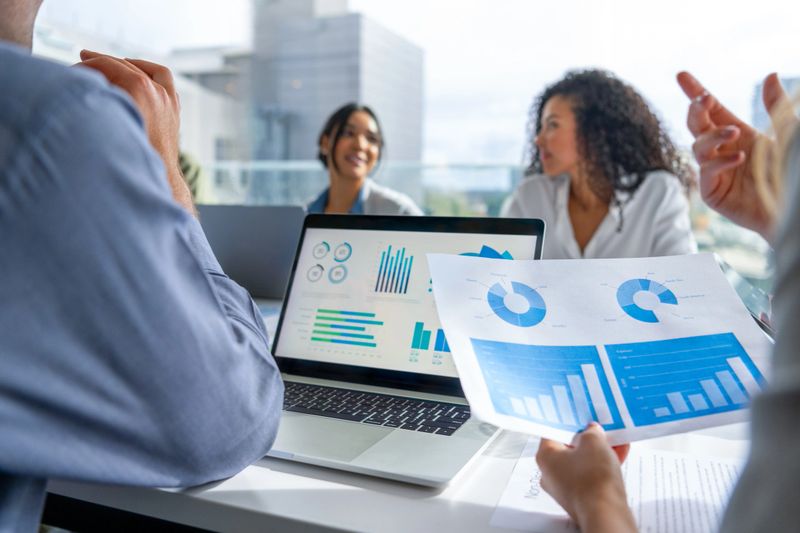 Close up of a laptop and document with data and finance charts and graphs. Group of business people in a meeting in then board room in the background. There are laptops and other technology on the table along with documents showing financial data