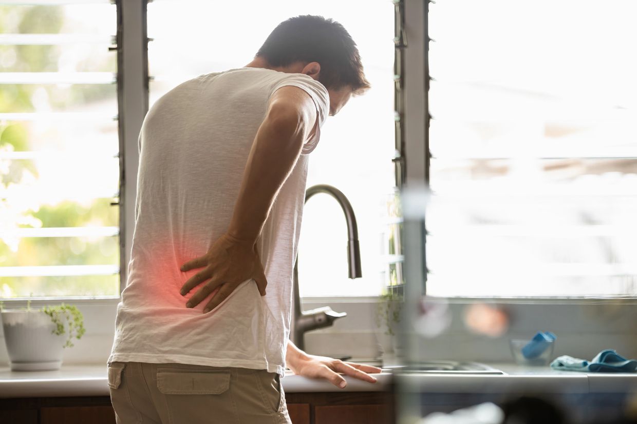 Man holding his lower back in pain in a bright kitchen.