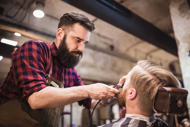 Man with a beard trimming the beard of a client in a modern barbershop during the daytime.