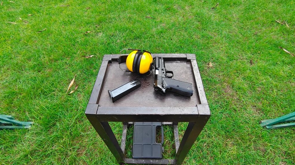A handgun, magazine, and yellow ear protection on a small wooden table outdoors.