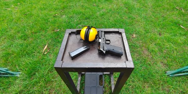 A handgun, magazine, and yellow ear protection on a wooden table outdoors.