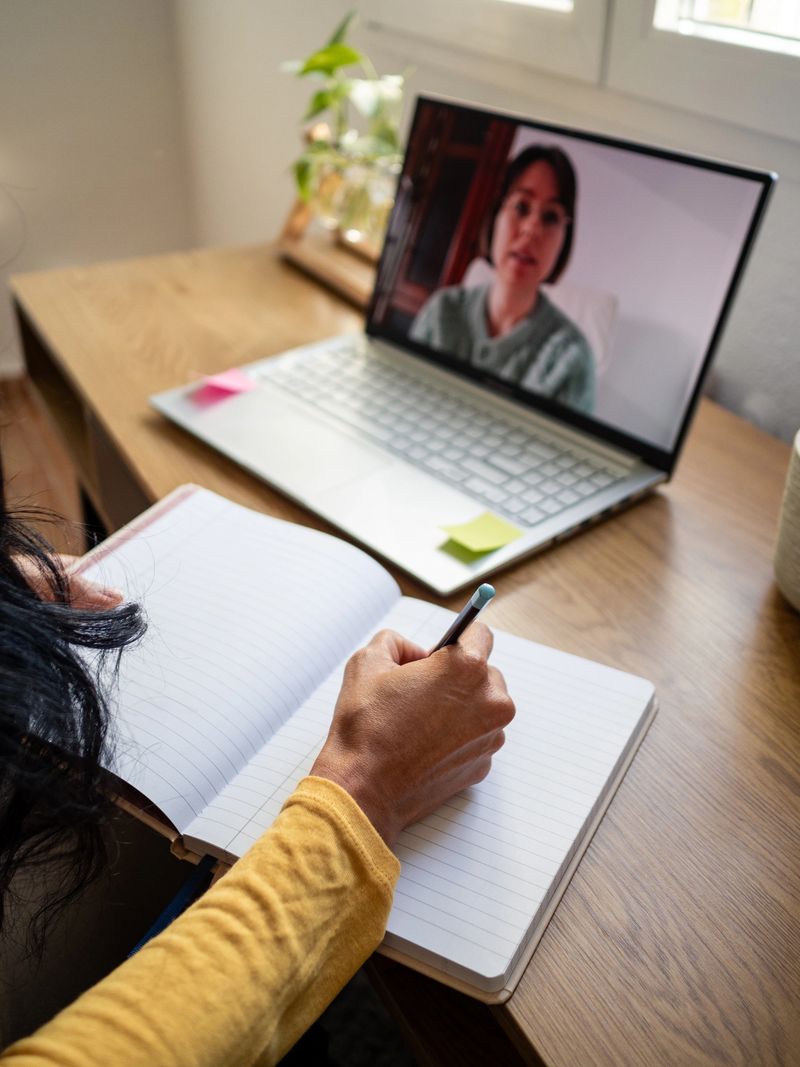 Hand of a Latin woman taking notes in a notebook while conversing at an online coaching session. The setting is a warm, cozy home office.