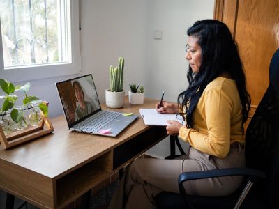 Woman attending a video call, taking notes at a wooden desk.