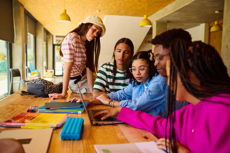 Diverse group of high school students working together on a school project, using a laptop in the library