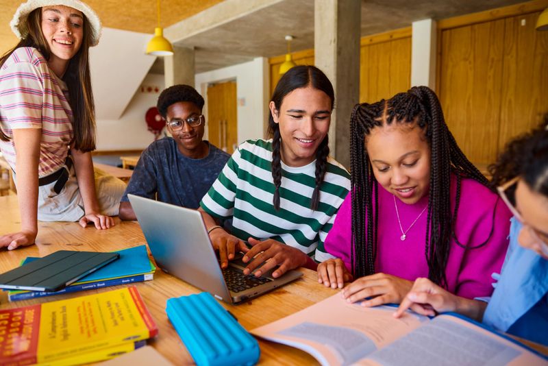 Diverse group of high school students working together on a school project, using a laptop in the library