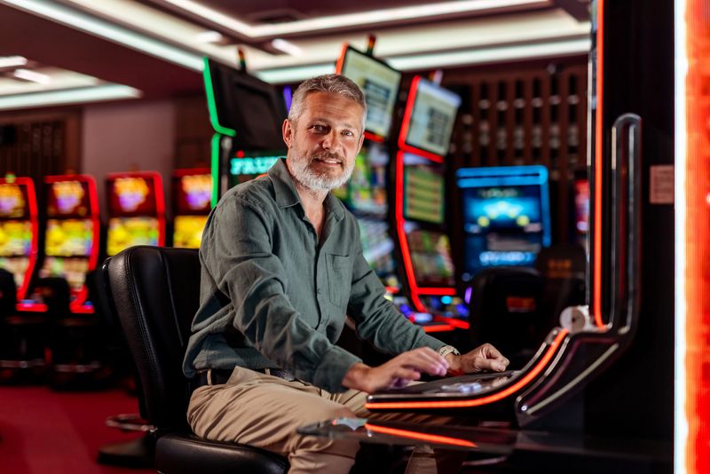 A mature man sits confidently at a brightly lit casino slot machine, surrounded by glowing screens. The vibrant atmosphere conveys excitement and entertainment while highlighting the sophisticated allure of gambling.