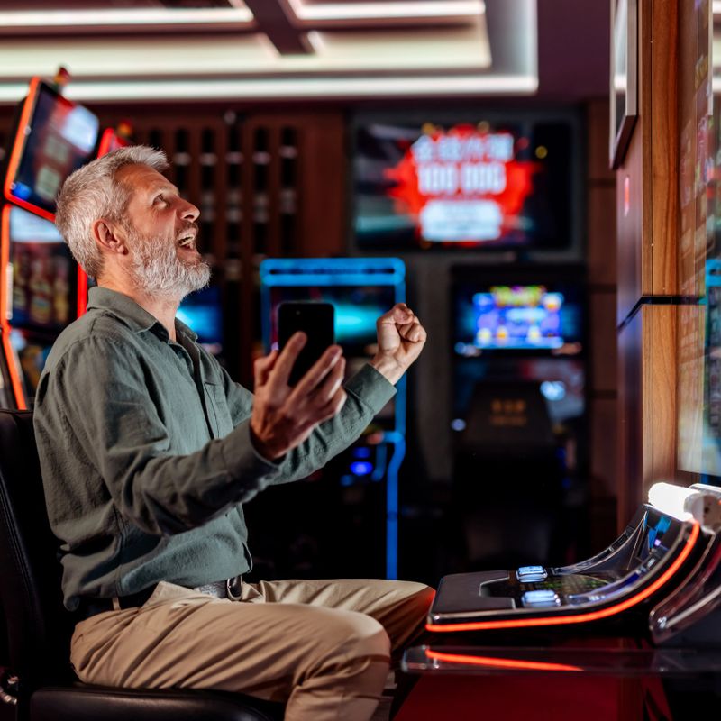 An older man celebrates winning while playing a slot machine in a vibrant casino setting, expressing excitement.