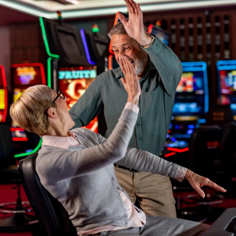 An excited senior couple engages cheerfully in a victory celebration while seated at a casino with glowing slot machines in the background, expressing joy and accomplishment.