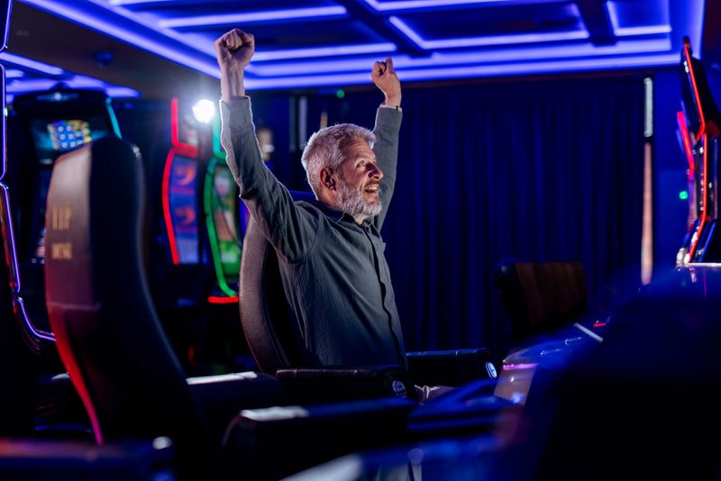 A cheerful elderly gentleman triumphantly raises his hands in celebration at a casino. The vibrant lights and slot machines convey excitement and achievement in this dynamic gaming setting.