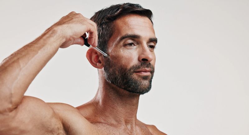 Hands, man and serum for skincare in studio for beard oil, facial treatment and dermatology cosmetics. Person, product and collagen for hair growth, hyaluronic acid and anti aging on white background