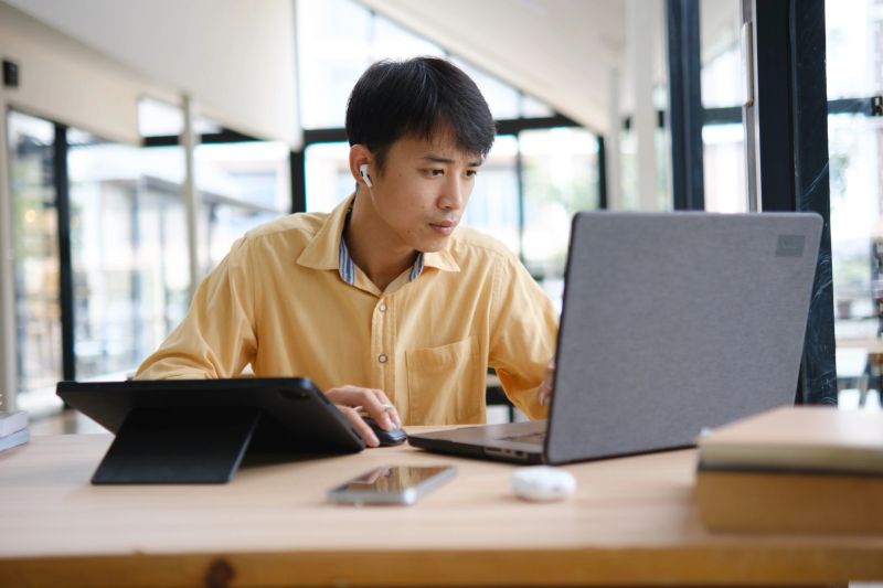 A student engaged in online learning, taking notes and reviewing study materials with books and digital devices to prepare for exams.