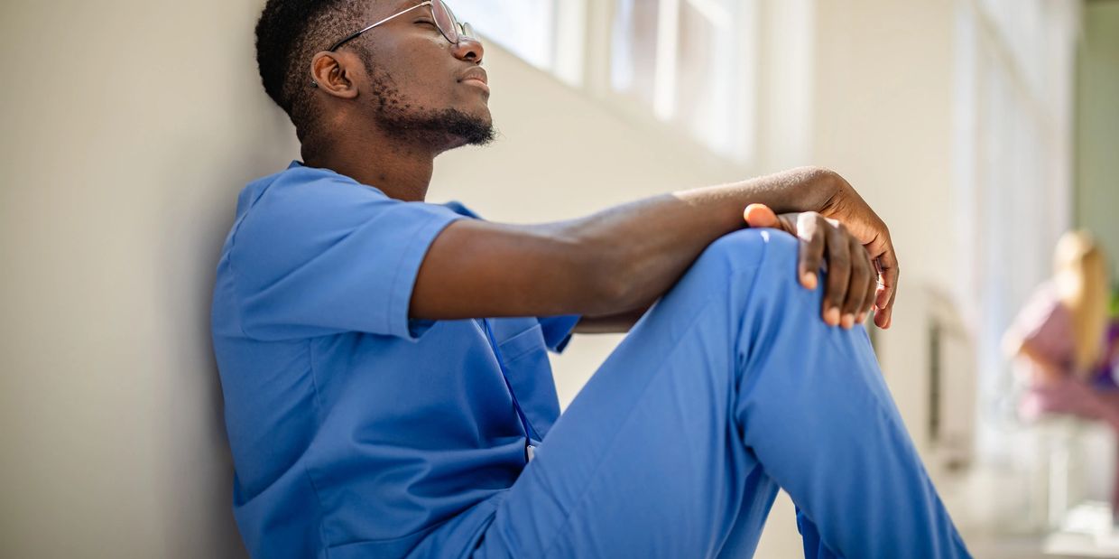 Tired healthcare worker in blue scrubs resting against a wall.