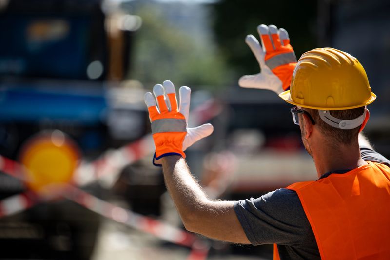 A construction worker in safety gear is directing vehicles at a construction site. Bright orange gloves and a helmet stand out against the background of equipment and barriers.