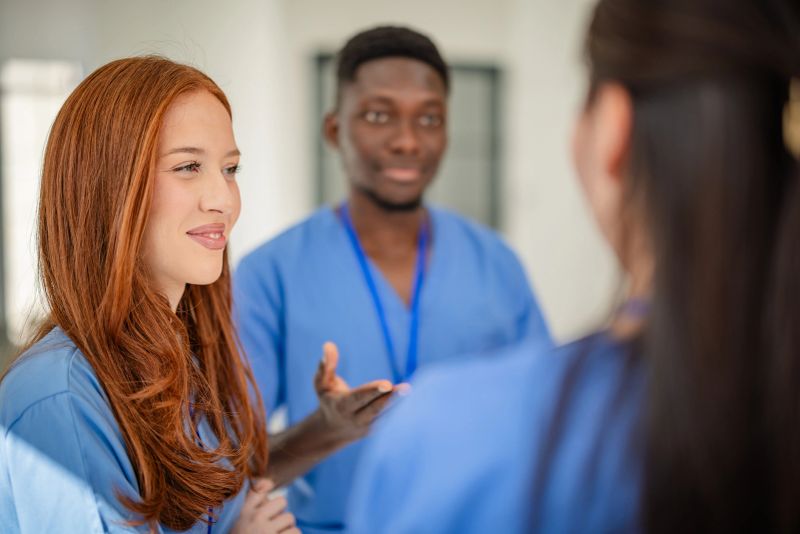 A group of young healthcare workers converses in a hospital. They are wearing medical scrubs and discussing plans and insights, showcasing teamwork in a professional environment.