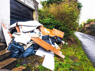Piled-up trash and discarded cardboard near an old garage door by a roadside.