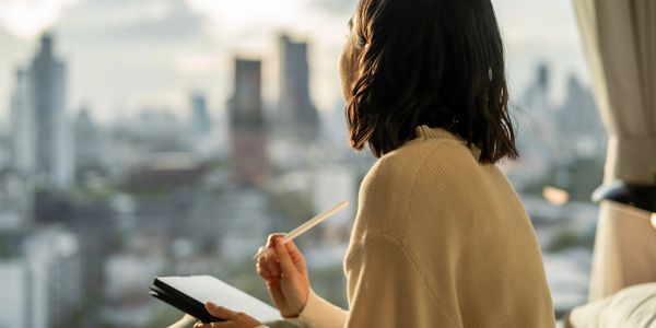 Woman in cozy sweater holding a tablet and stylus, gazing out at a cityscape.