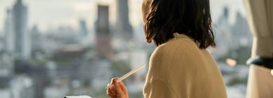 Person writing in a notebook while overlooking a cityscape from indoors.