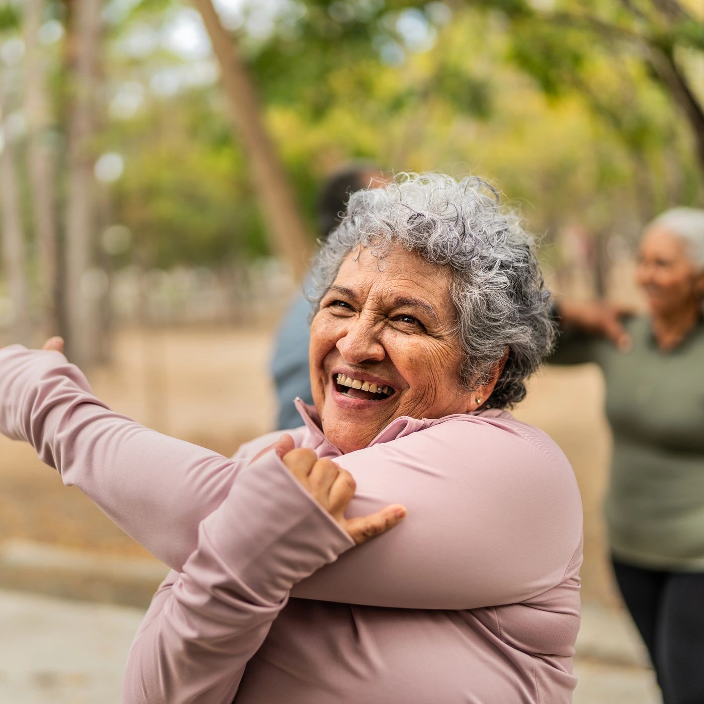 Smiling elderly woman stretching outdoors with friends in the background.