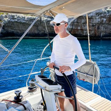 Man steering a sailboat on clear blue water near rocky cliffs.