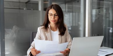 a woman with long dark hair, neutral color jacket, sitting & holding papers in a glass wall office