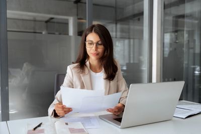 Businesswoman reviewing documents at her desk with a laptop.