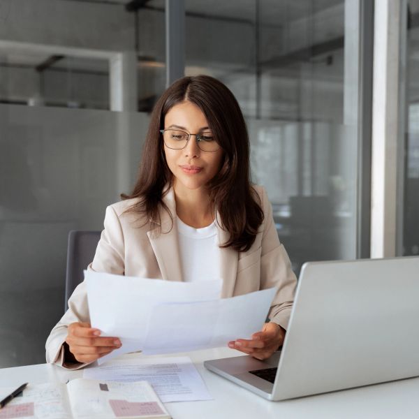 Woman reading documents at desk with laptop.