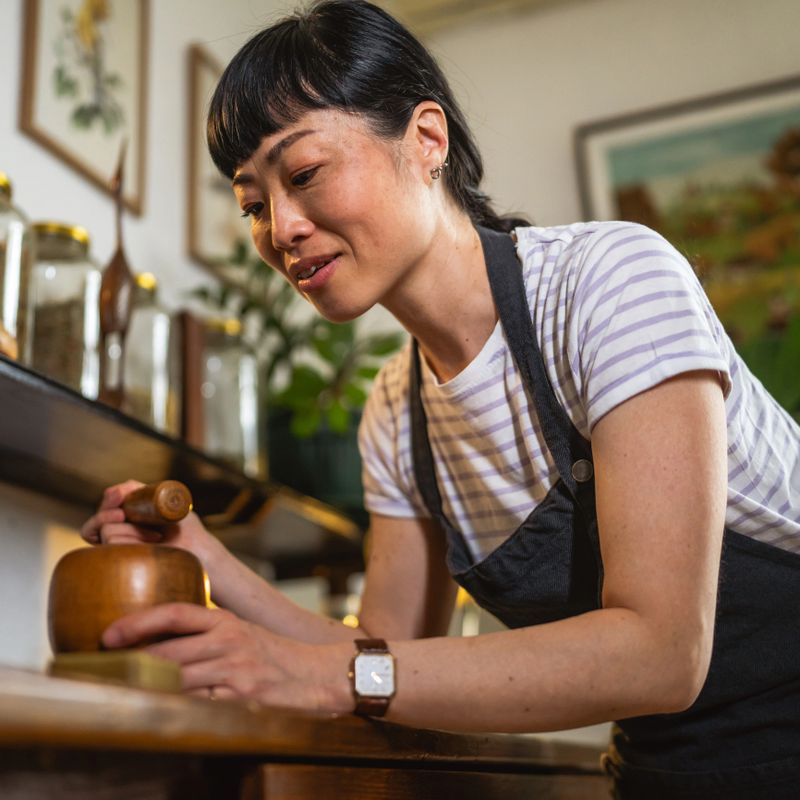 Mature japanese woman wear apron and use mortar and pestle on a wooden table to make soap in herbal workshop