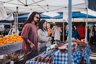 Couple shopping at a farmers market with fresh produce and a dog.