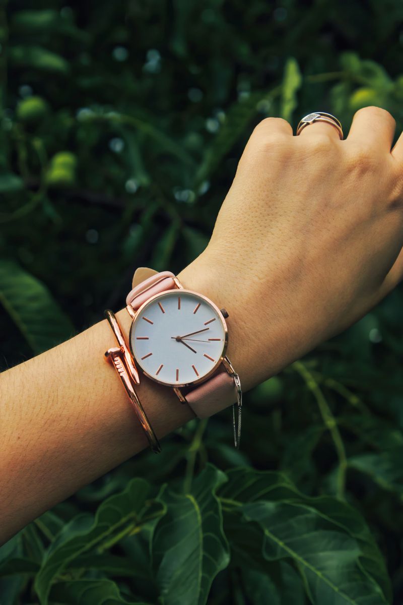 Close-up of wrist adorned with minimalist rose gold wristwatch and matching bracelet. White watch face contrasts elegantly with blurred green foliage background, emphasizing sophistication and style.