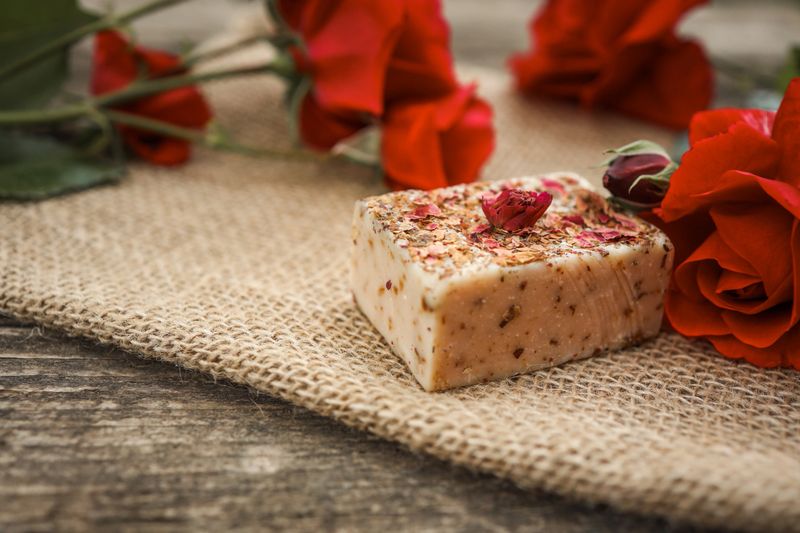 Handmade rose soap bar with dried petals on top, placed on burlap with fresh red roses. Natural skincare concept captured in a rustic, elegant setting.