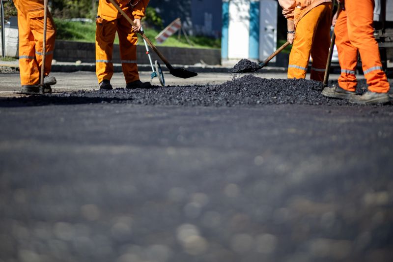 Construction workers in bright orange uniforms spread freshly laid asphalt on a road. They use shovels to ensure an even surface. The setting is sunny with minimal traffic disruption.