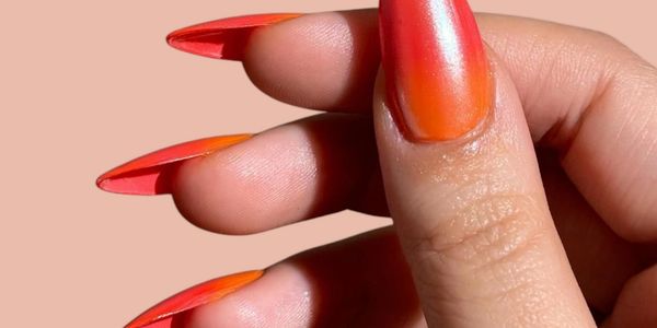 Hand with sharp, ombre orange to red stiletto nails against a peach background.