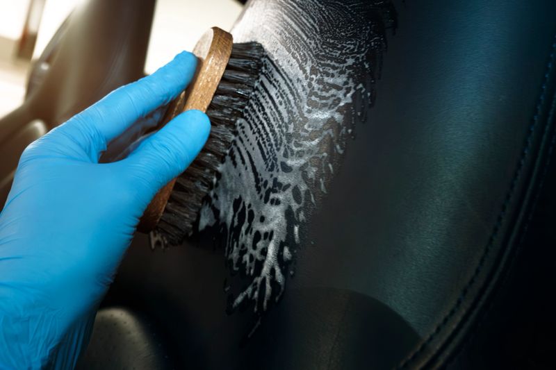 Hand Wearing a Blue Glove Scrubs a Black Car Seat With a Brush, Applying Cleaning Solution to Maintain the Car’s Interior. The Focus Is on Cleanliness and Precision.