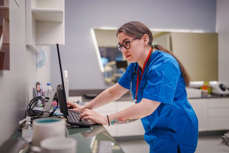 A female healthcare professional efficiently manages patient appointments, using both a computer and a mobile phone at a clinic counter. The scene highlights multitasking, technology use, and administrative duties in a medical environment.