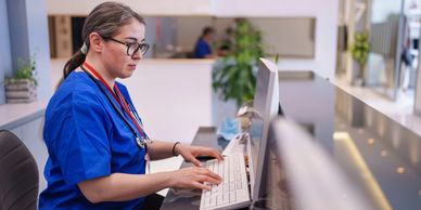 A female healthcare professional in blue scrubs working on a computer at a reception desk.